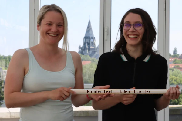zwei Frauen halten den Staffelstanb mit der Aufschrift "Runder Tisch – Talent Transfer"