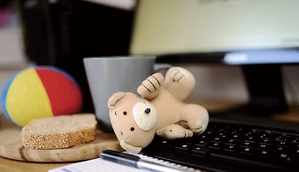 Plüsch-Teddybär auf einer Computer-Tastatur mit , Monitor, Tasse, Brot und Ball im Hintergrund